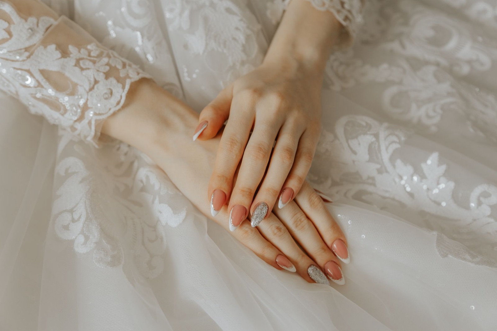 Handen van een bruid met amandelvormige nude nagels met een witte French-rand en zilverglitter, op een kanten trouwjurk