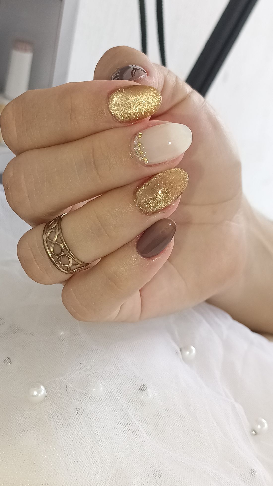 Hand met nagels in goudglitter, crème en chocoladebruin, met een gouden ring