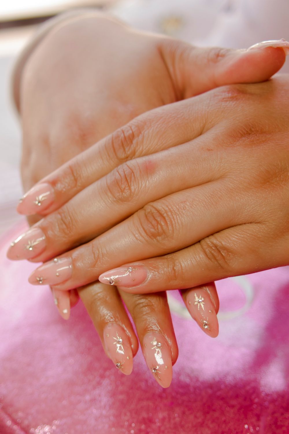 Handen met lange, amandelvormige nude nagels met kleine gouden details op roze glinsterstof