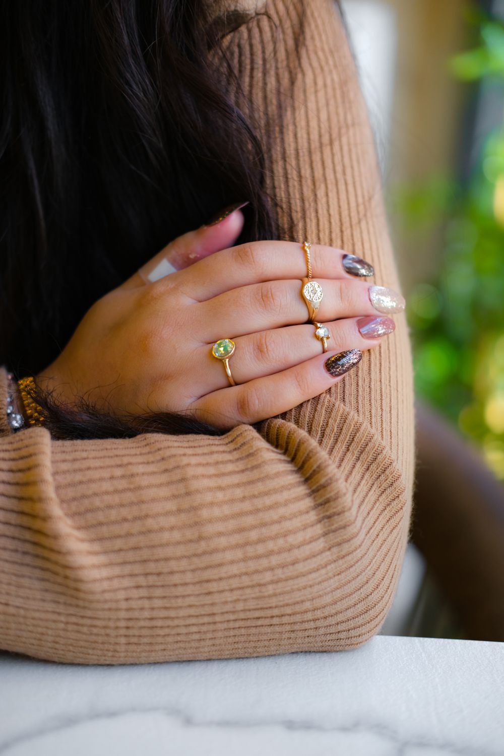 Hand met versierde nagels en gouden ringen op de mouw van een camelkleurige trui