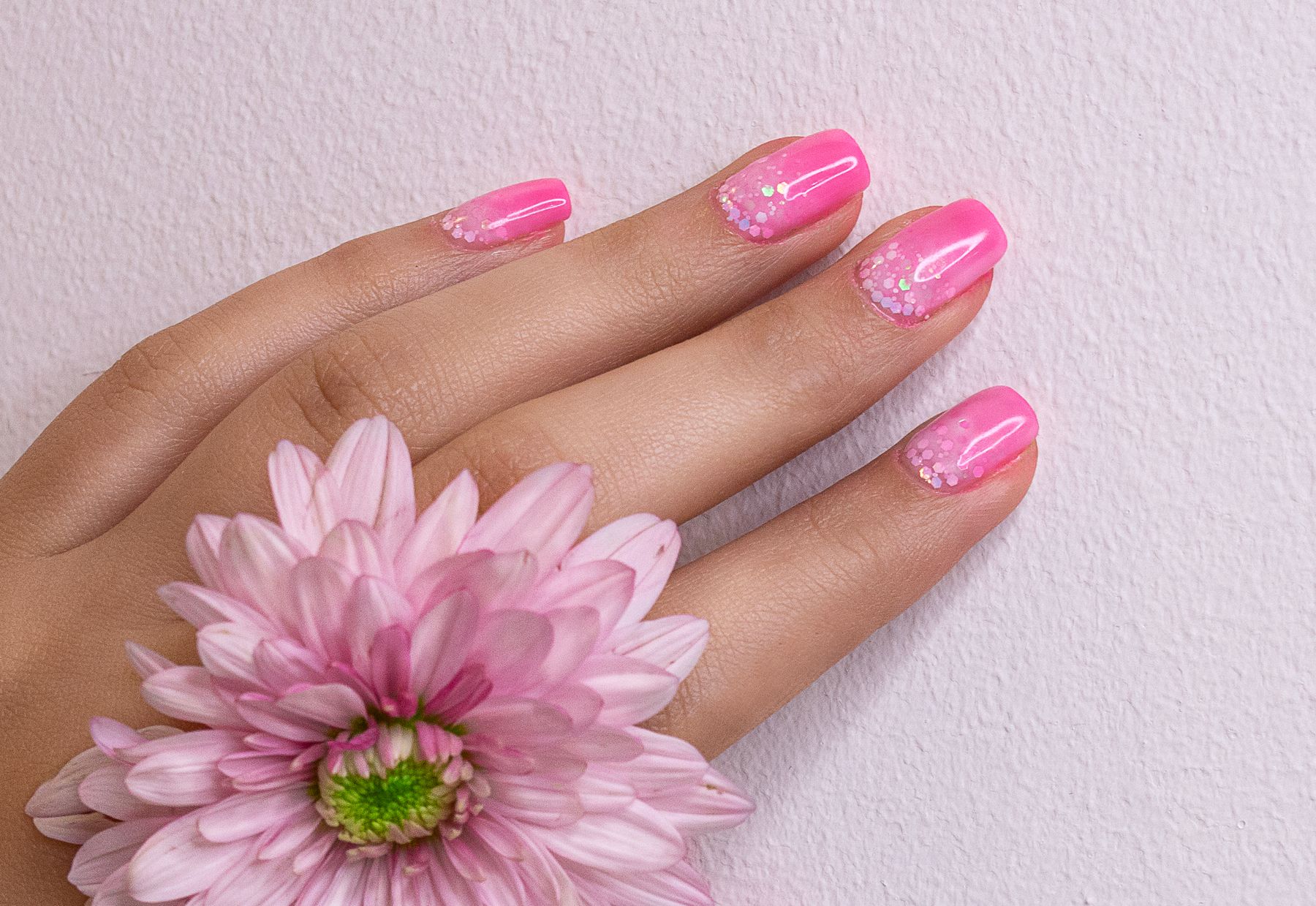 Hand met felroze nagels met fijne glitter en een roze bloem tegen een lichtroze achtergrond