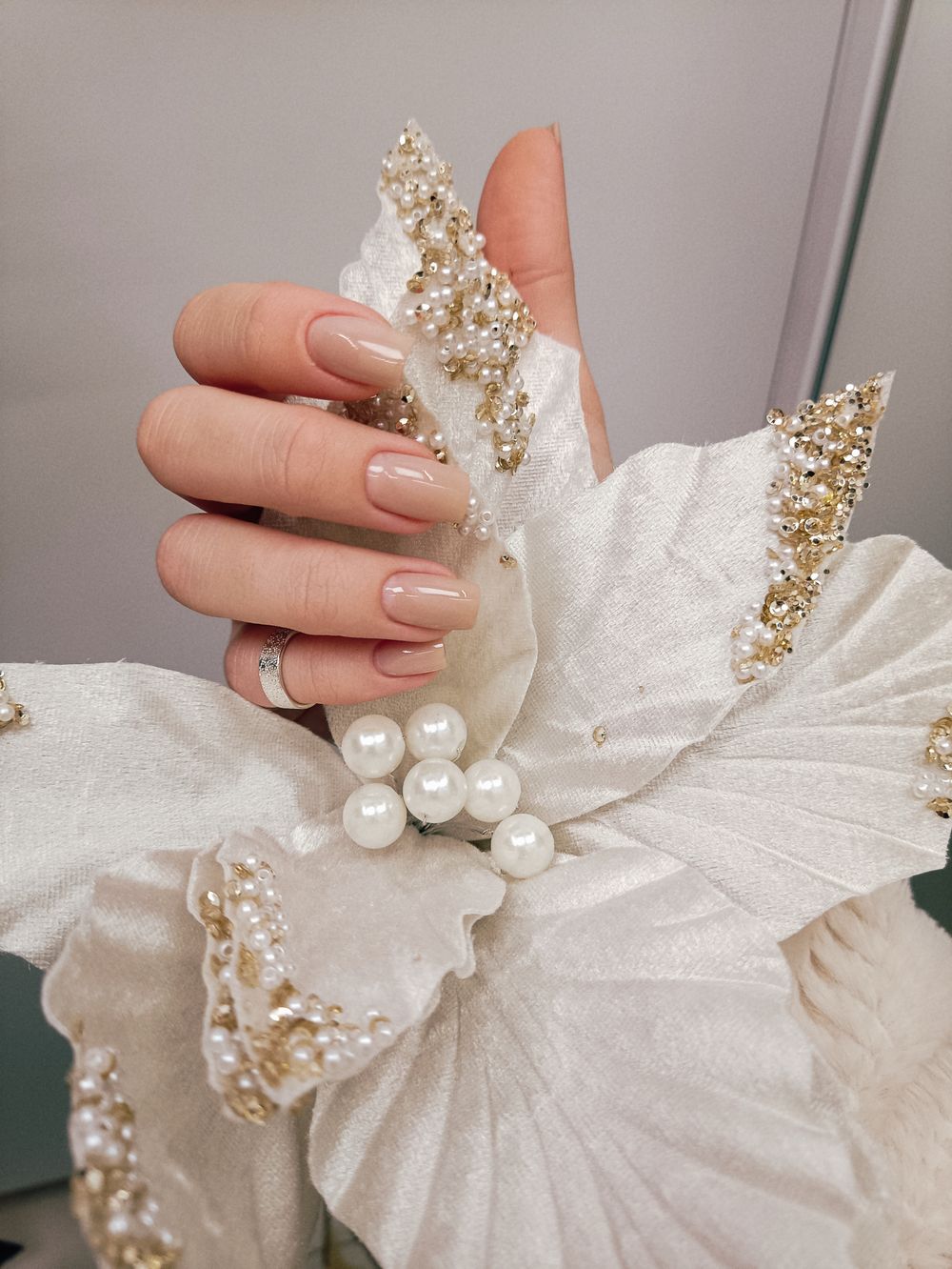 Hand met nude nagels boven een witte sierbloem met parels en goudglitter